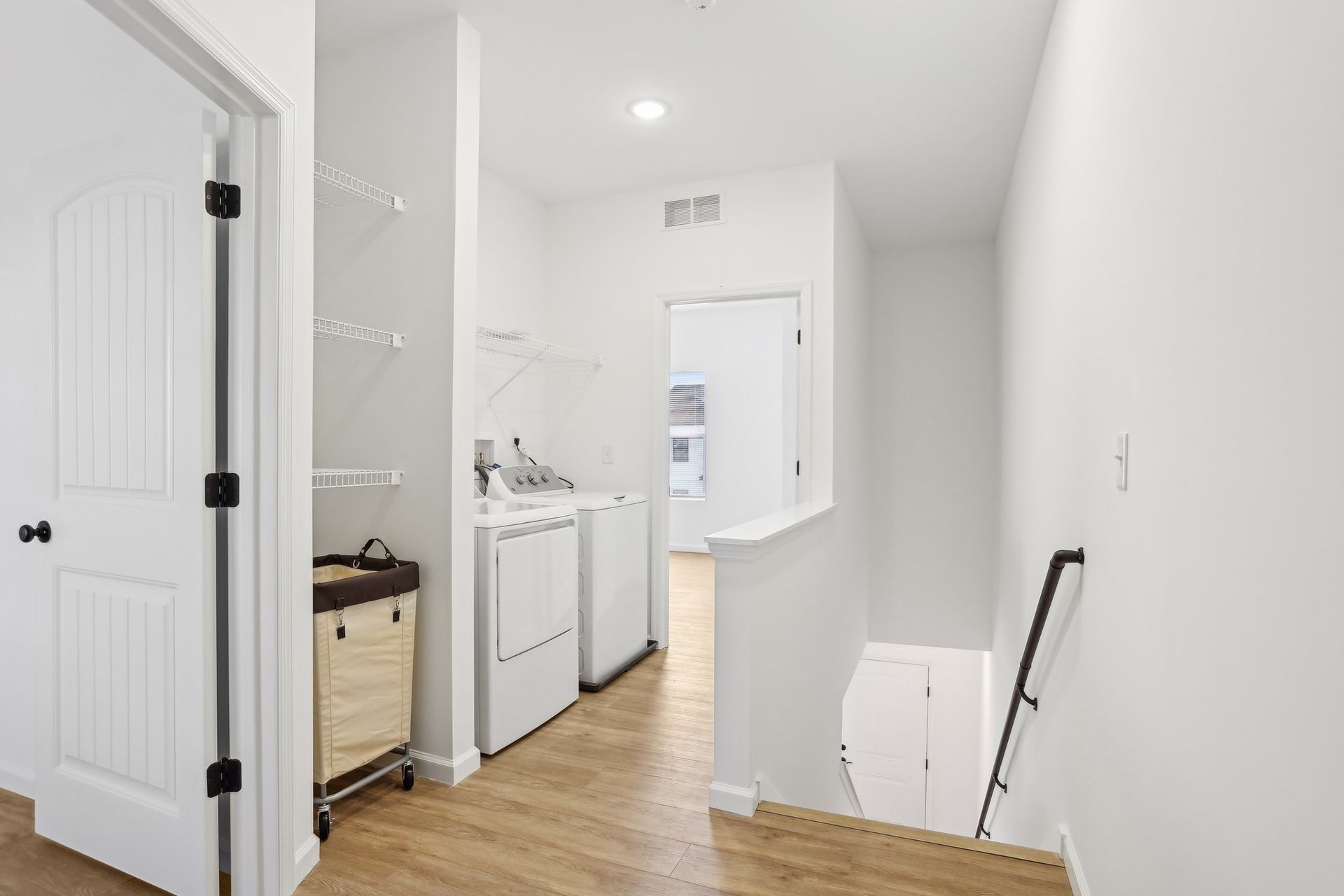Bright white hallway with a small laundry nook, open doors, wood floor, and a stair railing on the right