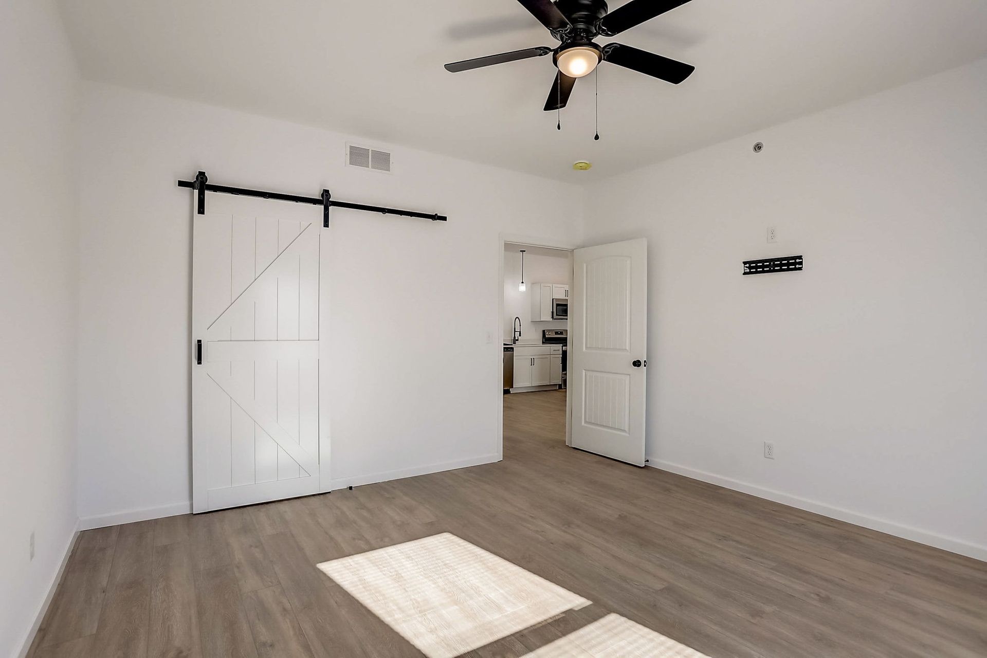 Empty white room with gray wood floors, barn-style sliding door, ceiling fan, and open doorway to a bathroom.