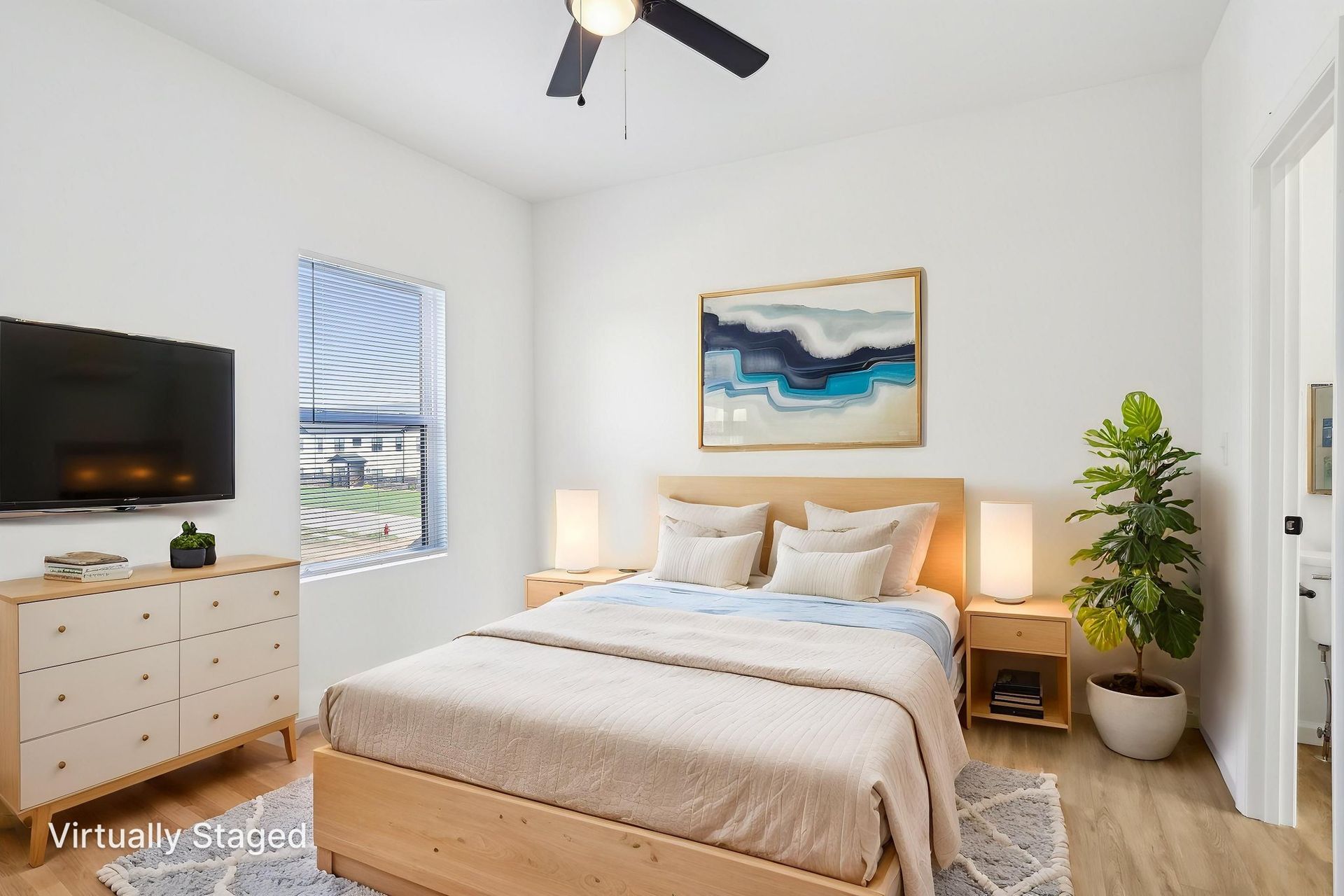 Bright bedroom with beige bed, white walls, wall art, TV, dresser, and potted plant by the window.