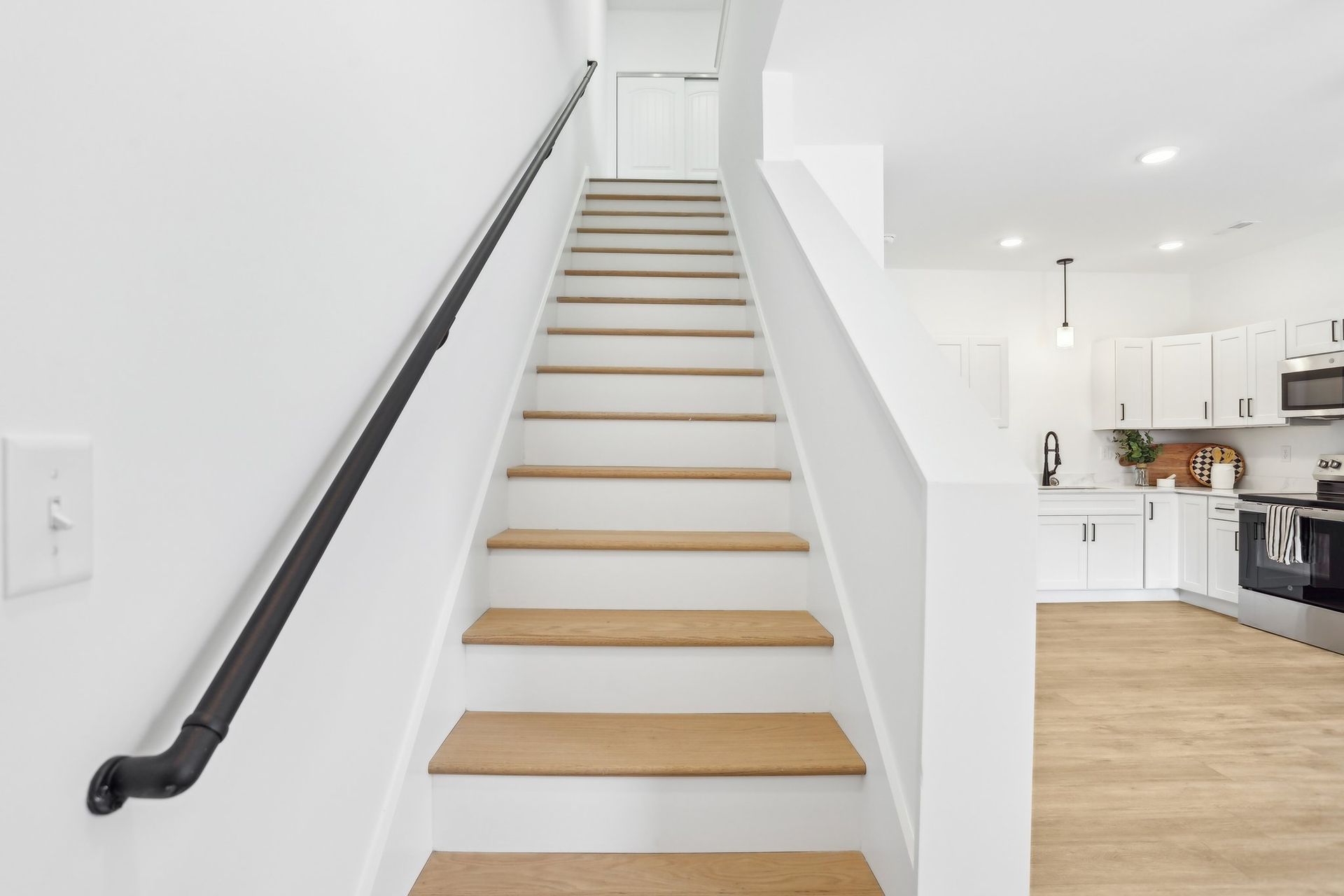 Bright modern staircase with white walls, light wood steps, and a black handrail leading to an open kitchen.