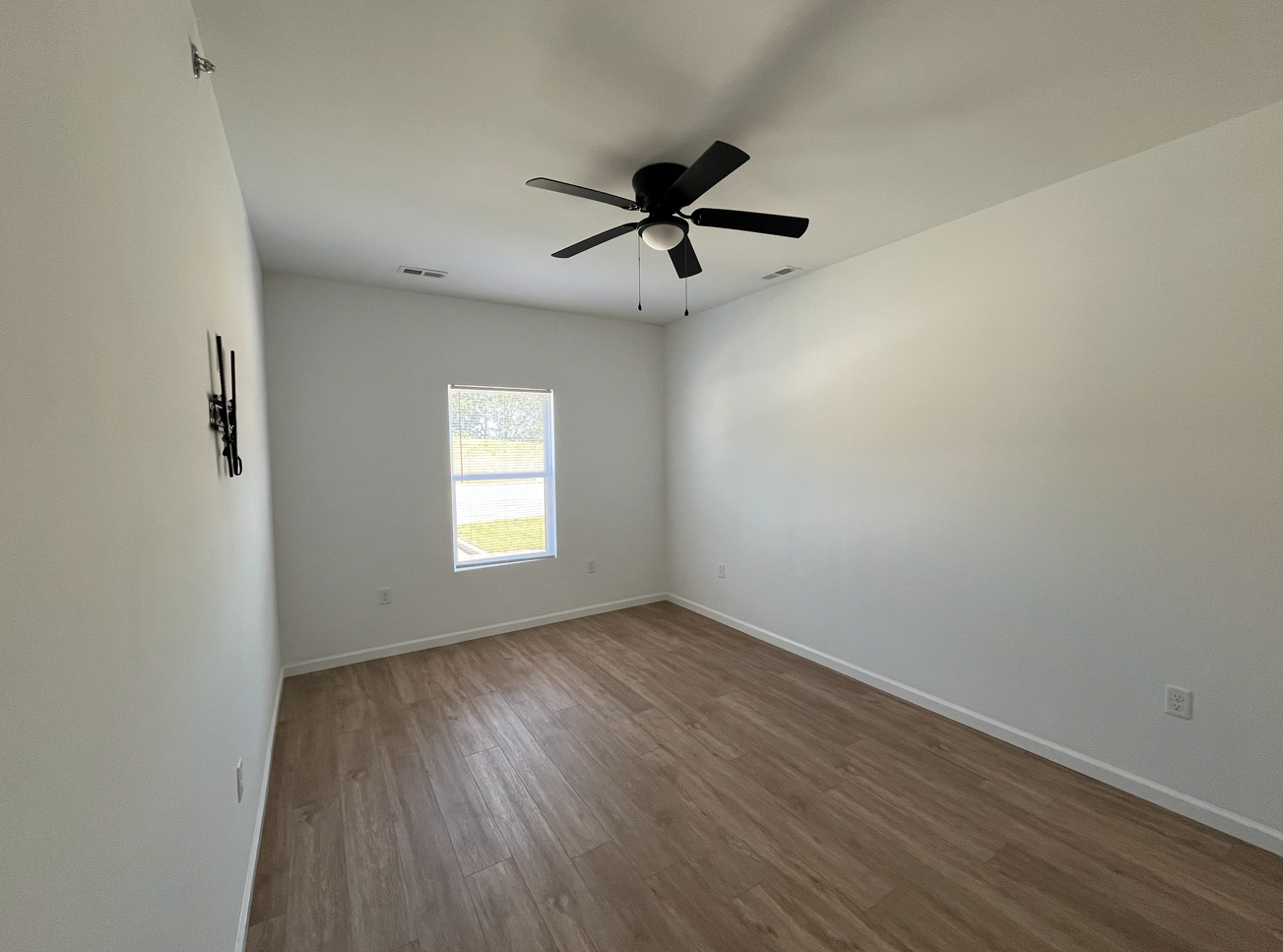 Empty white bedroom with wood flooring, a ceiling fan, and a small window.