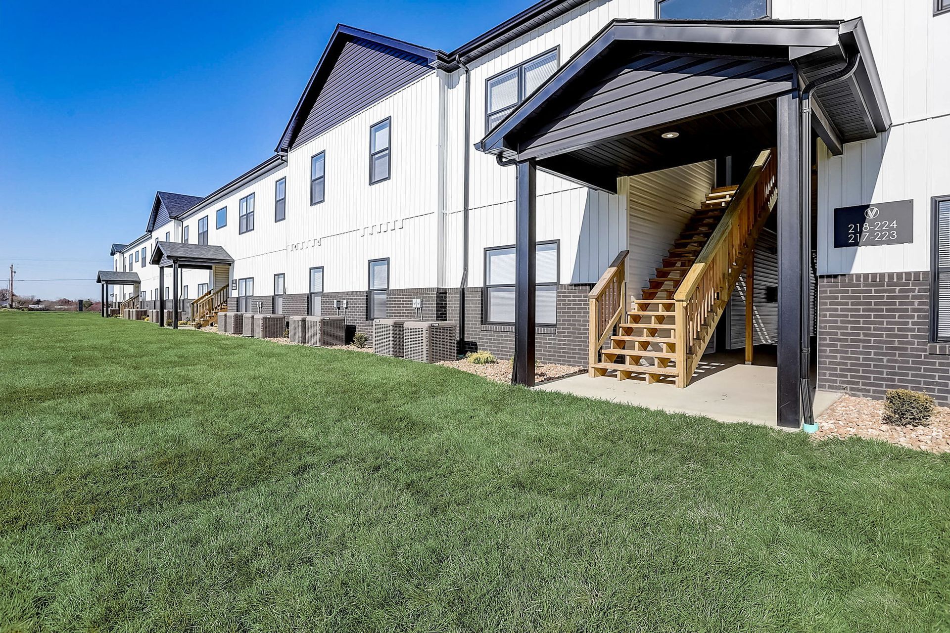 Apartment building exterior with covered staircase entrance and grassy lawn under a blue sky