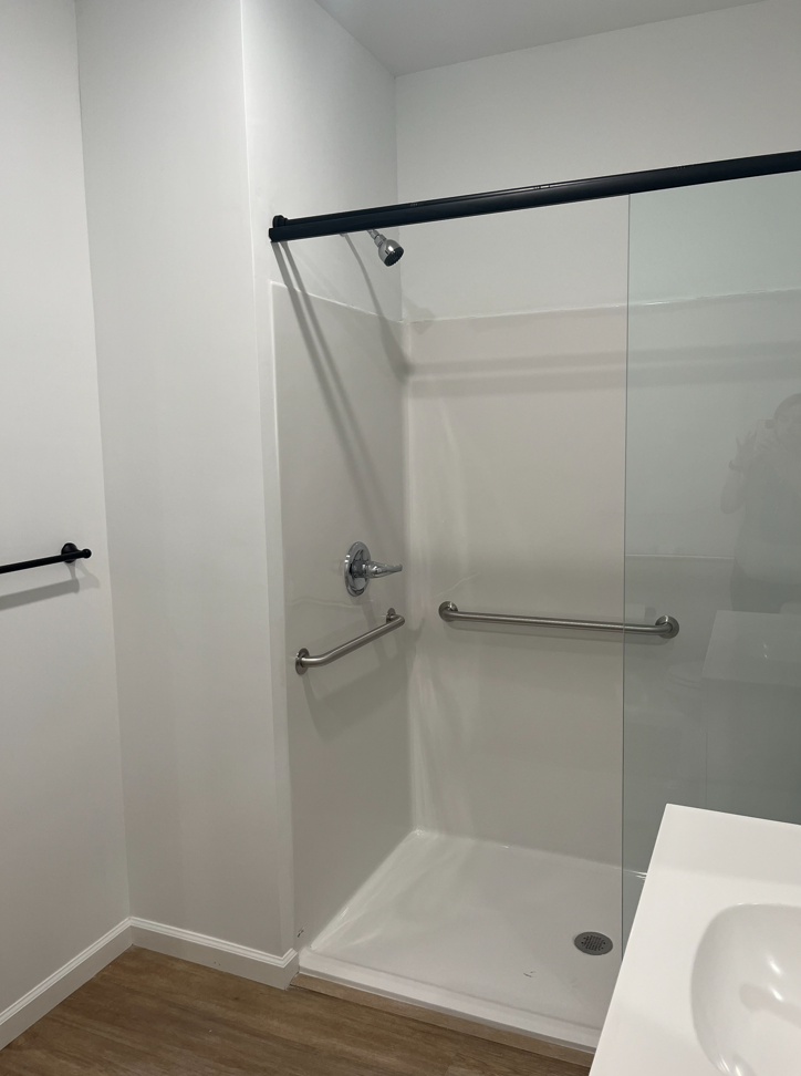 Accessible shower with black grab bars, clear glass panel, and a handheld showerhead in a white bathroom.