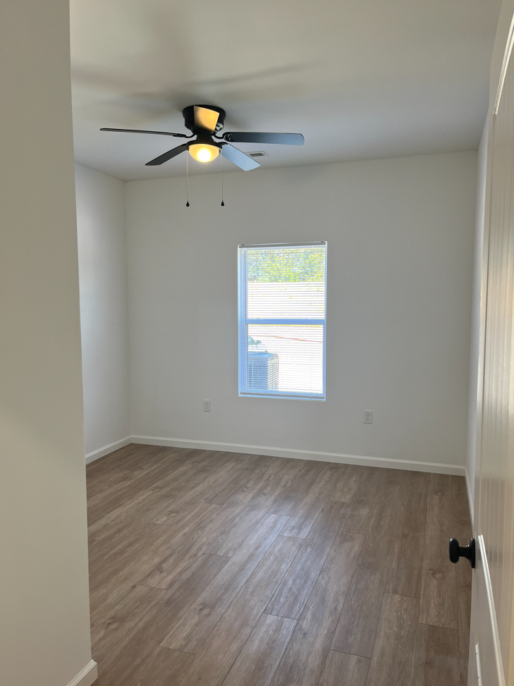 Empty small bedroom with wood floors, white walls, a ceiling fan, and a narrow window.