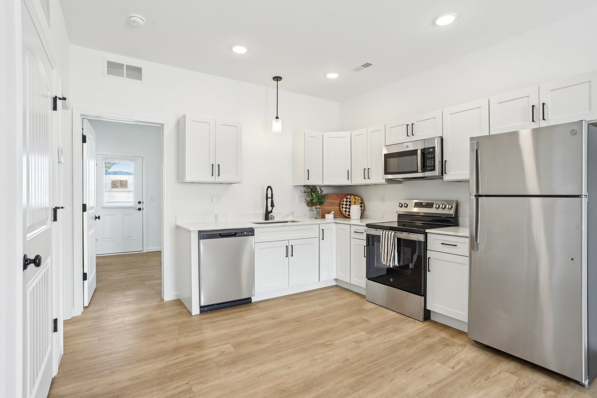 Bright white kitchen with stainless steel appliances, wood floors, and open doorway to another room