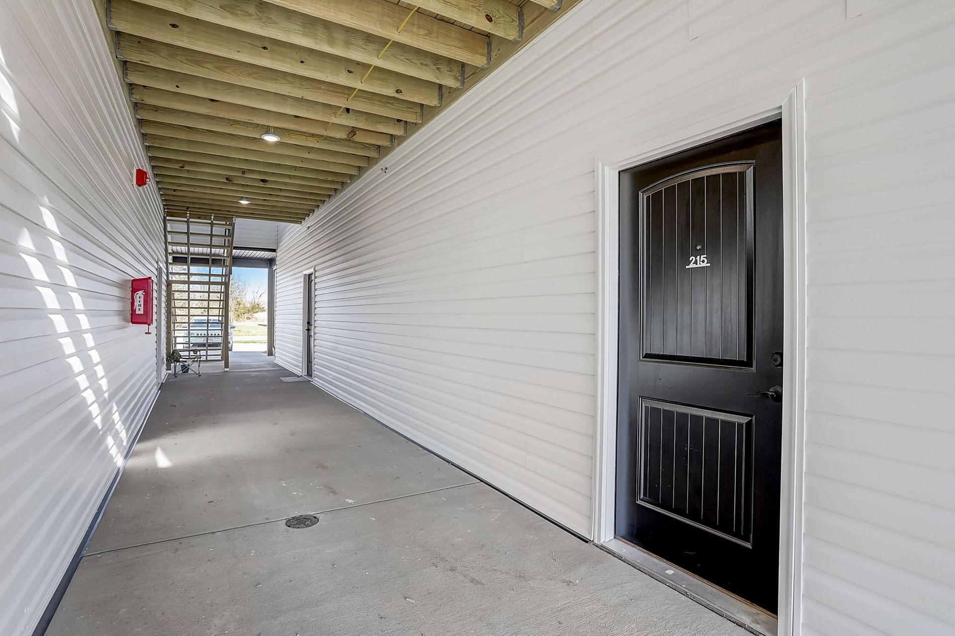 Covered outdoor corridor with white siding, black doors, and concrete walkway under a wood ceiling