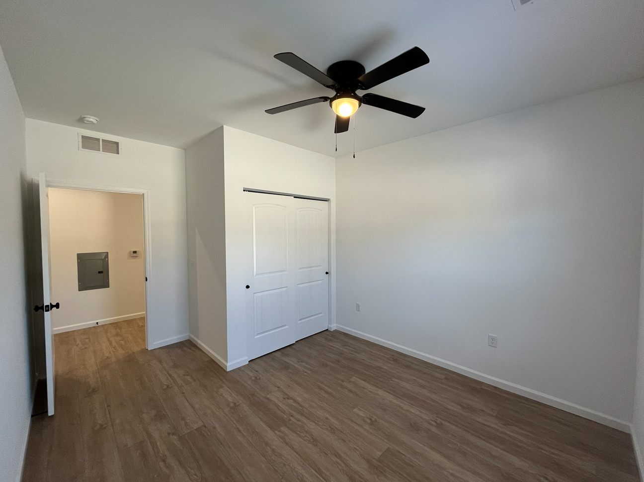 Empty bedroom with white walls, wood floor, ceiling fan, and closet doors