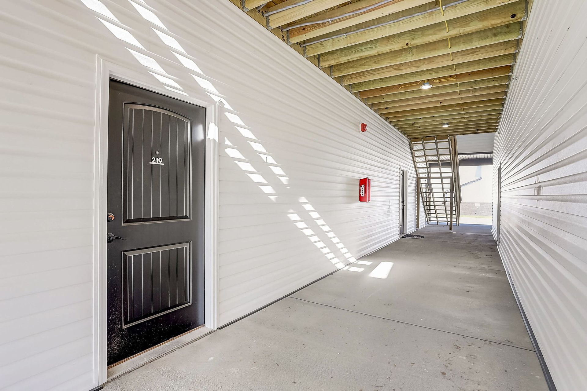 Covered outdoor walkway with white walls, concrete floor, black door, and stairs at the far end