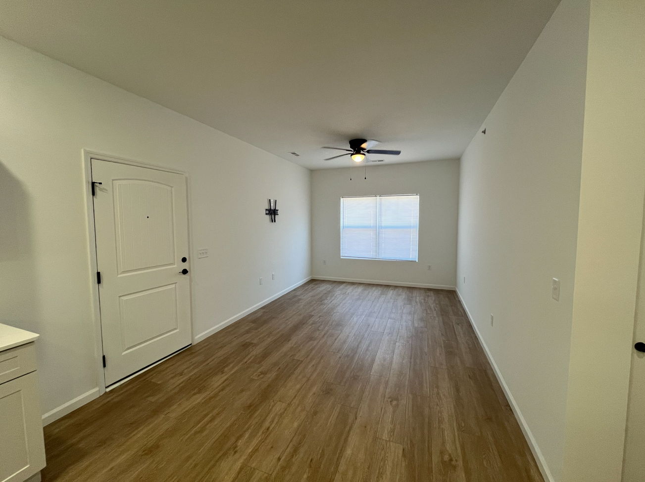 Empty white room with wood floors, a ceiling fan, and a window at the far end.