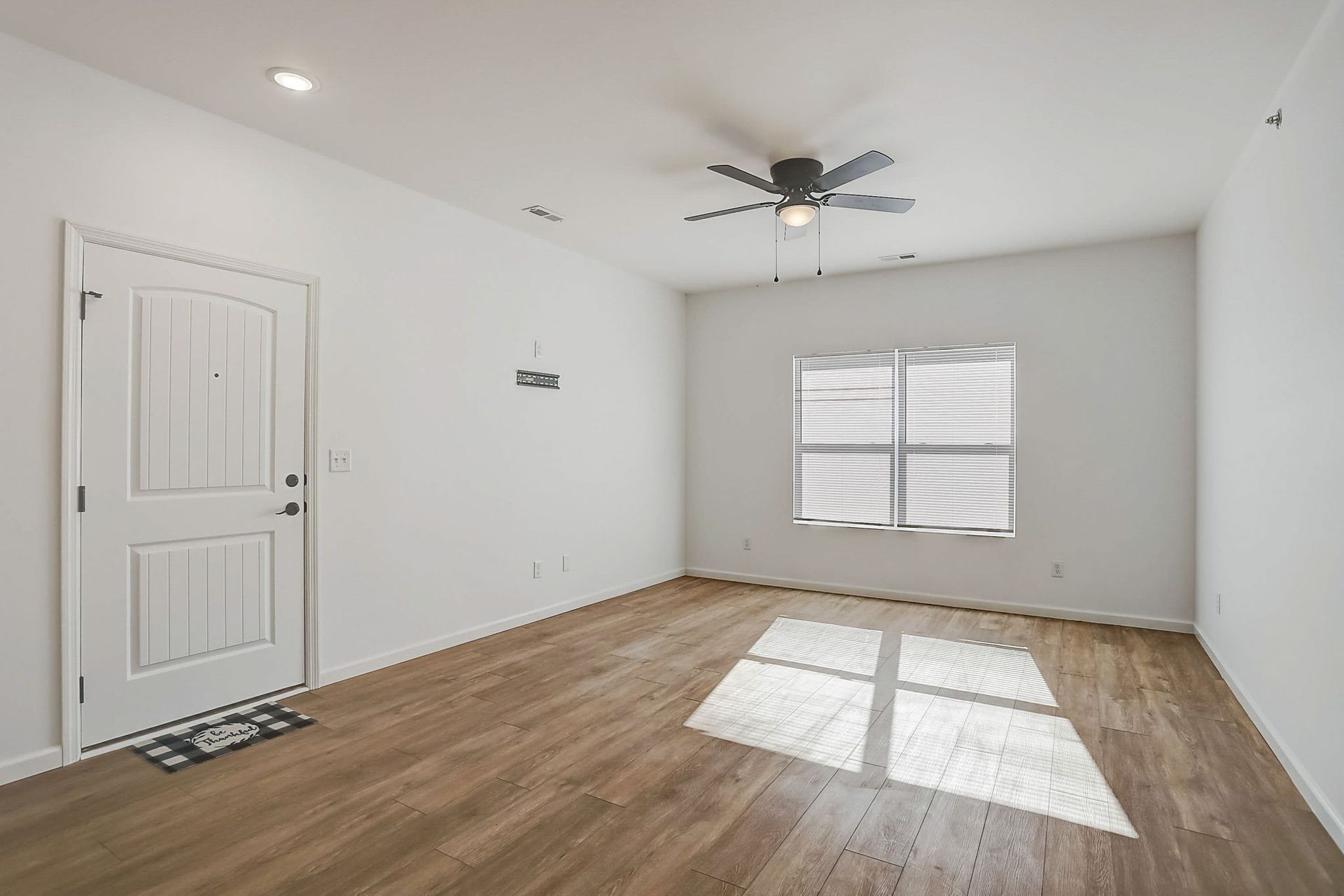 Empty white living room with wood floors, ceiling fan, and sunlight from a window