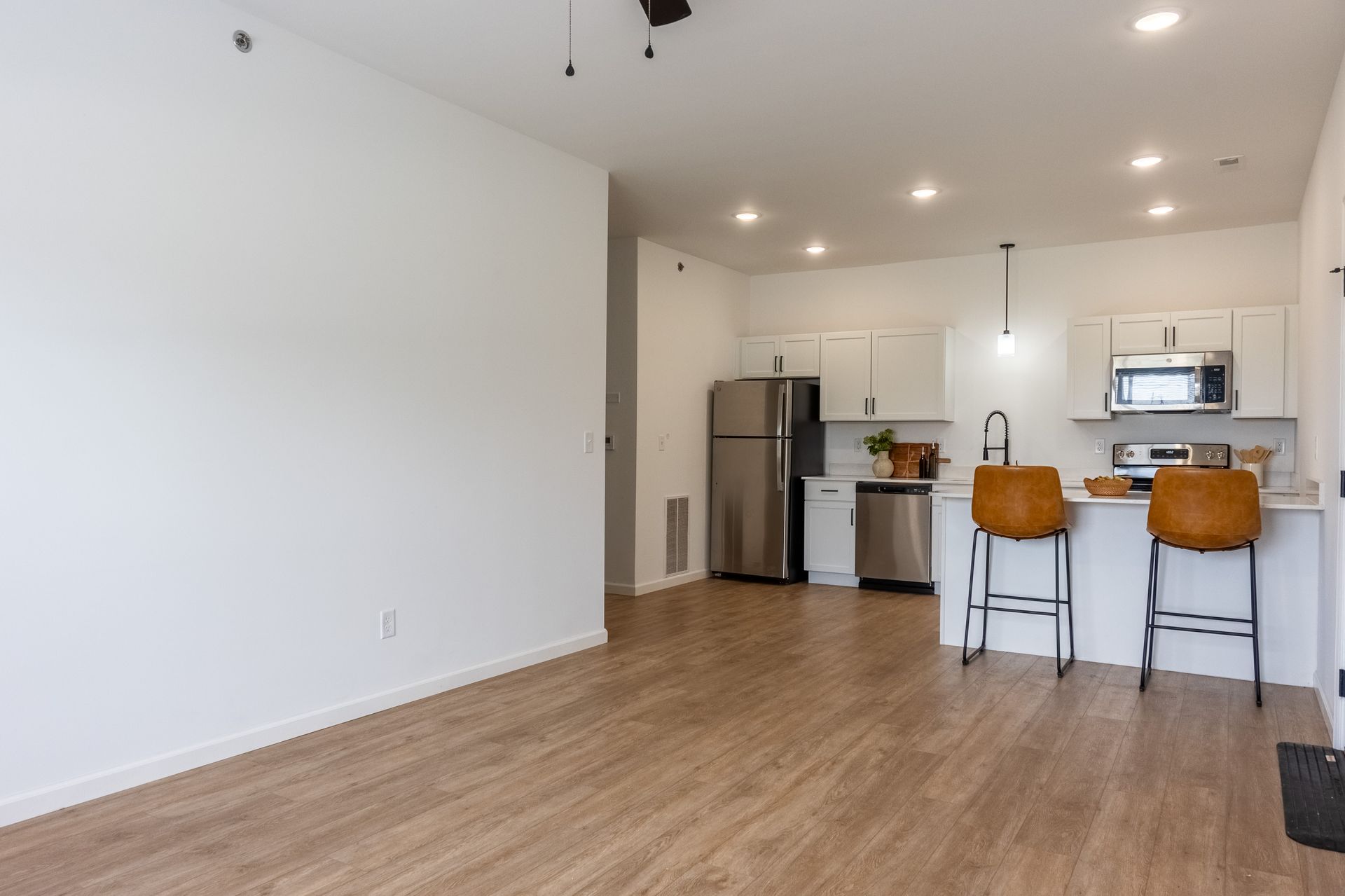 Empty modern apartment living area with wood floors, white walls, and a kitchen island with two stools.