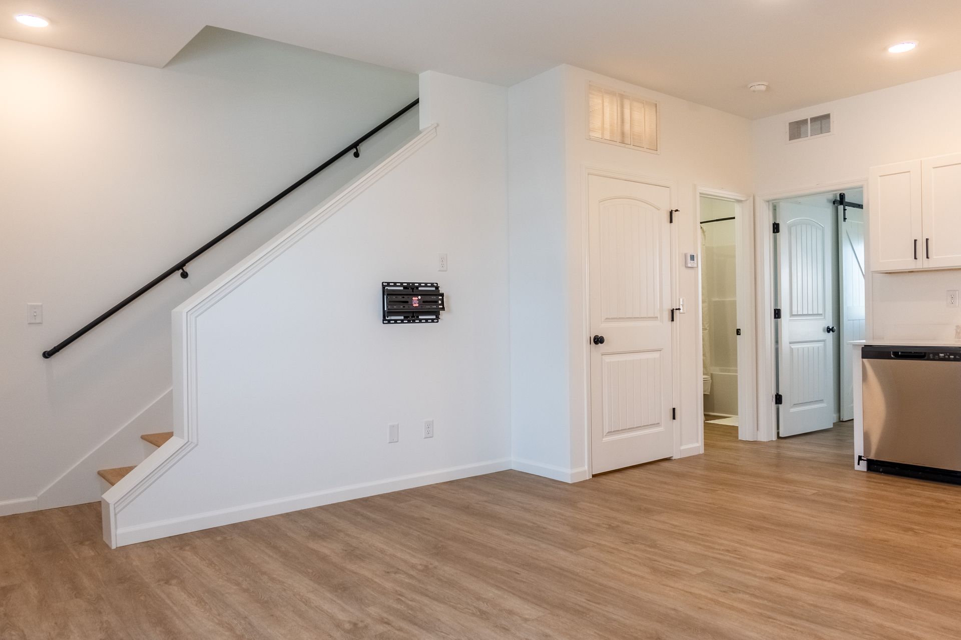 Bright empty room with staircase, white doors, light wood floors, and kitchen area.