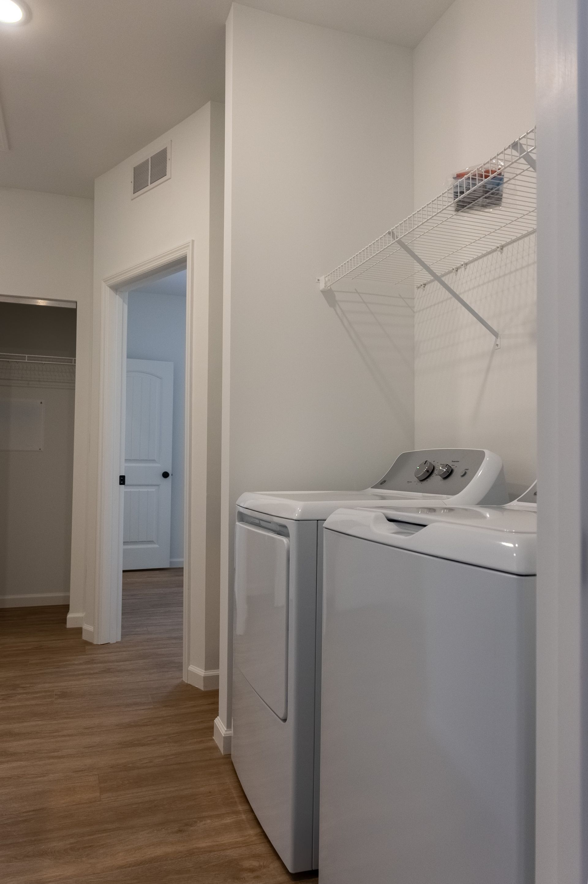 Laundry room with white washer and dryer, wire shelf, and hardwood hallway leading to a doorway