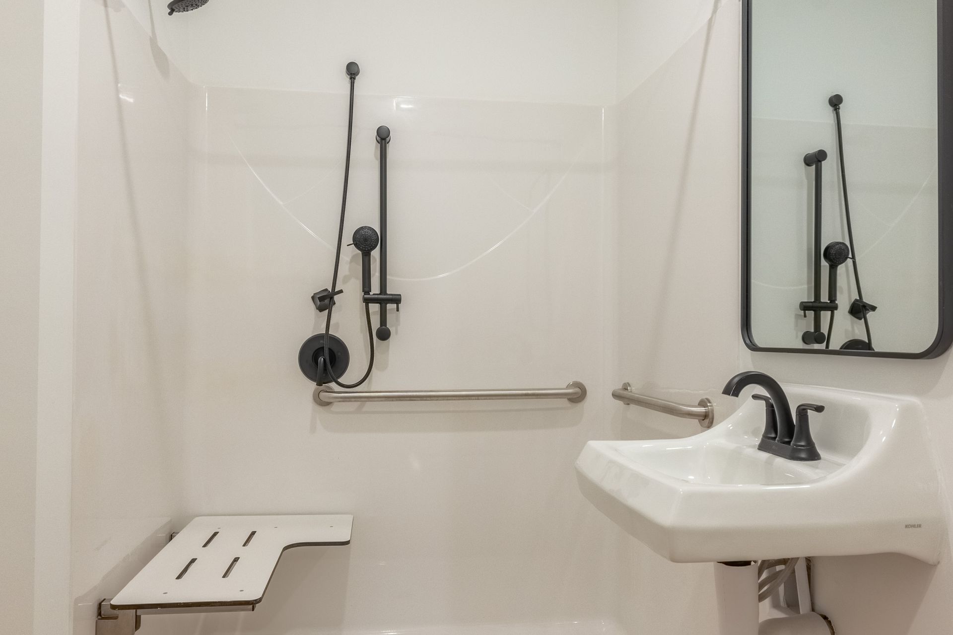 Accessible bathroom with shower, grab bars, shower seat, and sink beside a mirror.