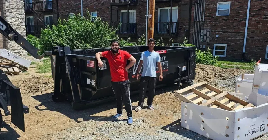 Two men are standing in front of a dumpster.