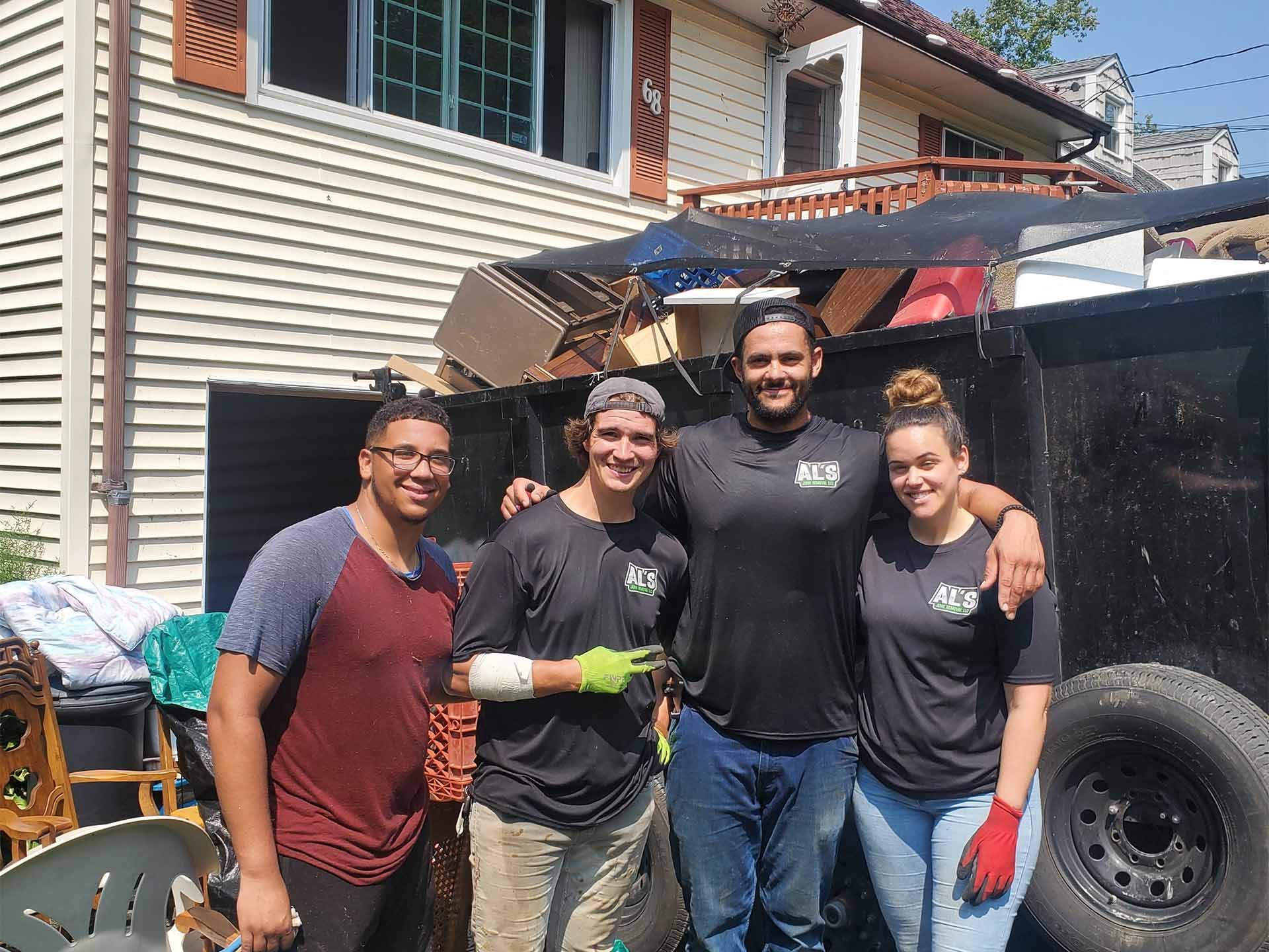 Four people standing in front of a trailer, likely after a cleanup. Items and a house are in the background.