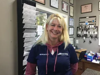 Smiling blonde woman in navy polo standing in an office with papers, certificates, and equipment on the walls | Pitts Automotive