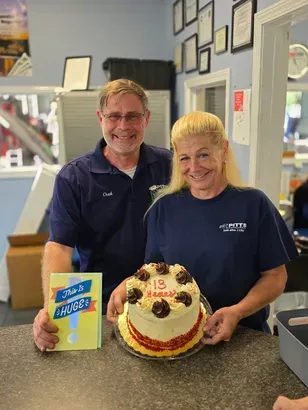 Two smiling coworkers pose with a decorated cake inside a shop, holding a colorful gift bag. | Pitts Automotive