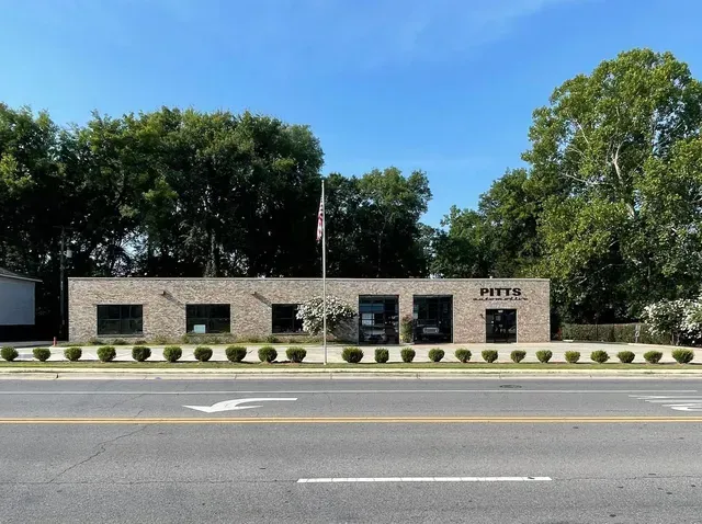 Stone building behind a roadside, lined with shrubs and trees under a blue sky | Pitts Automotive