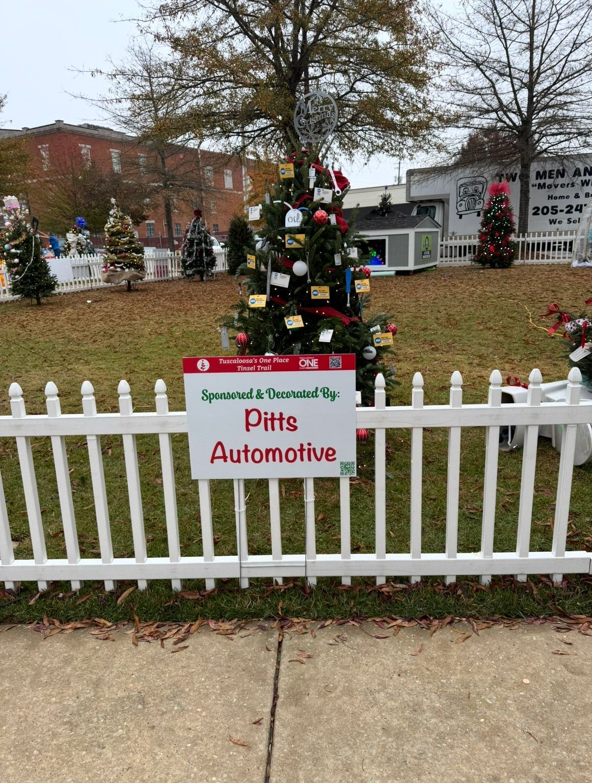 Holiday tree behind white fence with a Pitts Automotive sign in a grassy outdoor display | Pitts Automotive