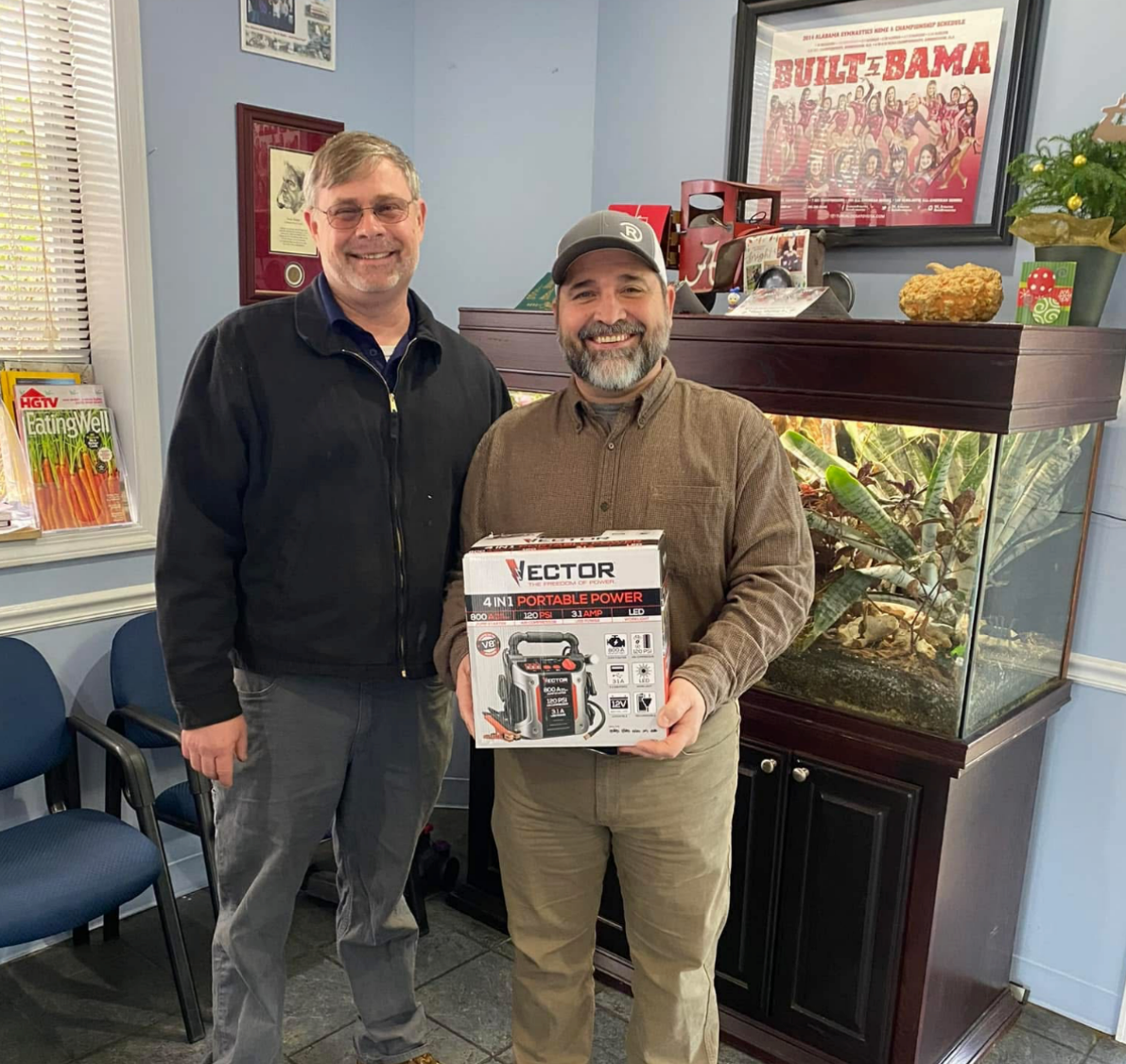 Two men smiling indoors, one holding a VEVOR box beside a fish tank and colorful wall art. | Pitts Automotive