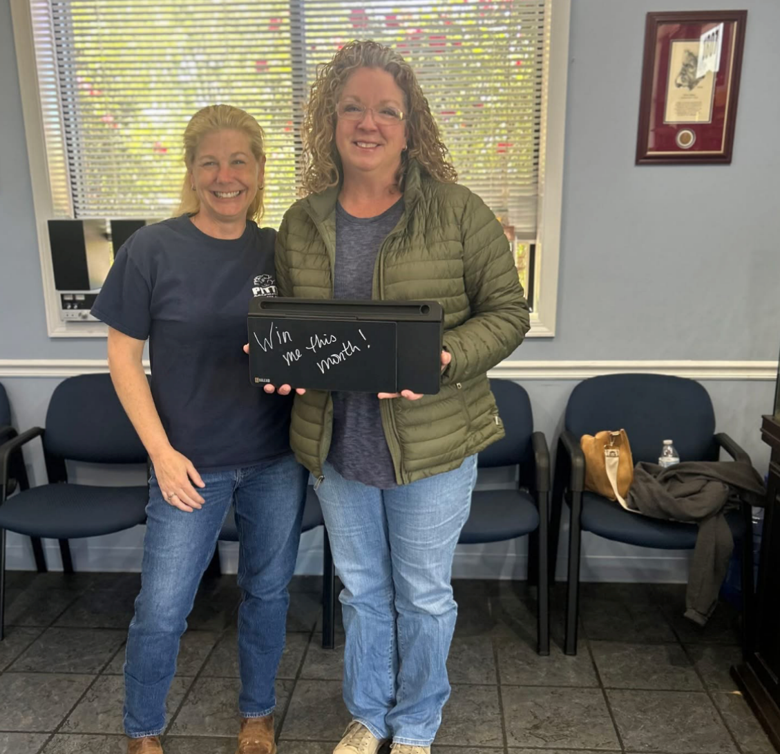 Two smiling people stand indoors holding a black box in front of chairs and a window. | Pitts Automotive