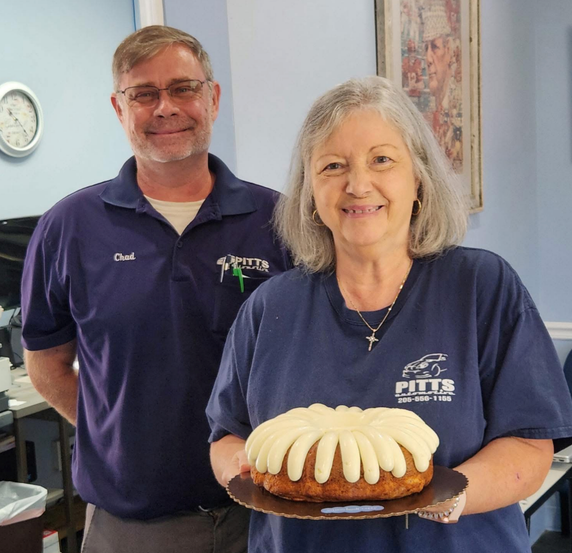 Two people in navy shirts holding a bundt cake indoors, one smiling in front. | Pitts Automotive