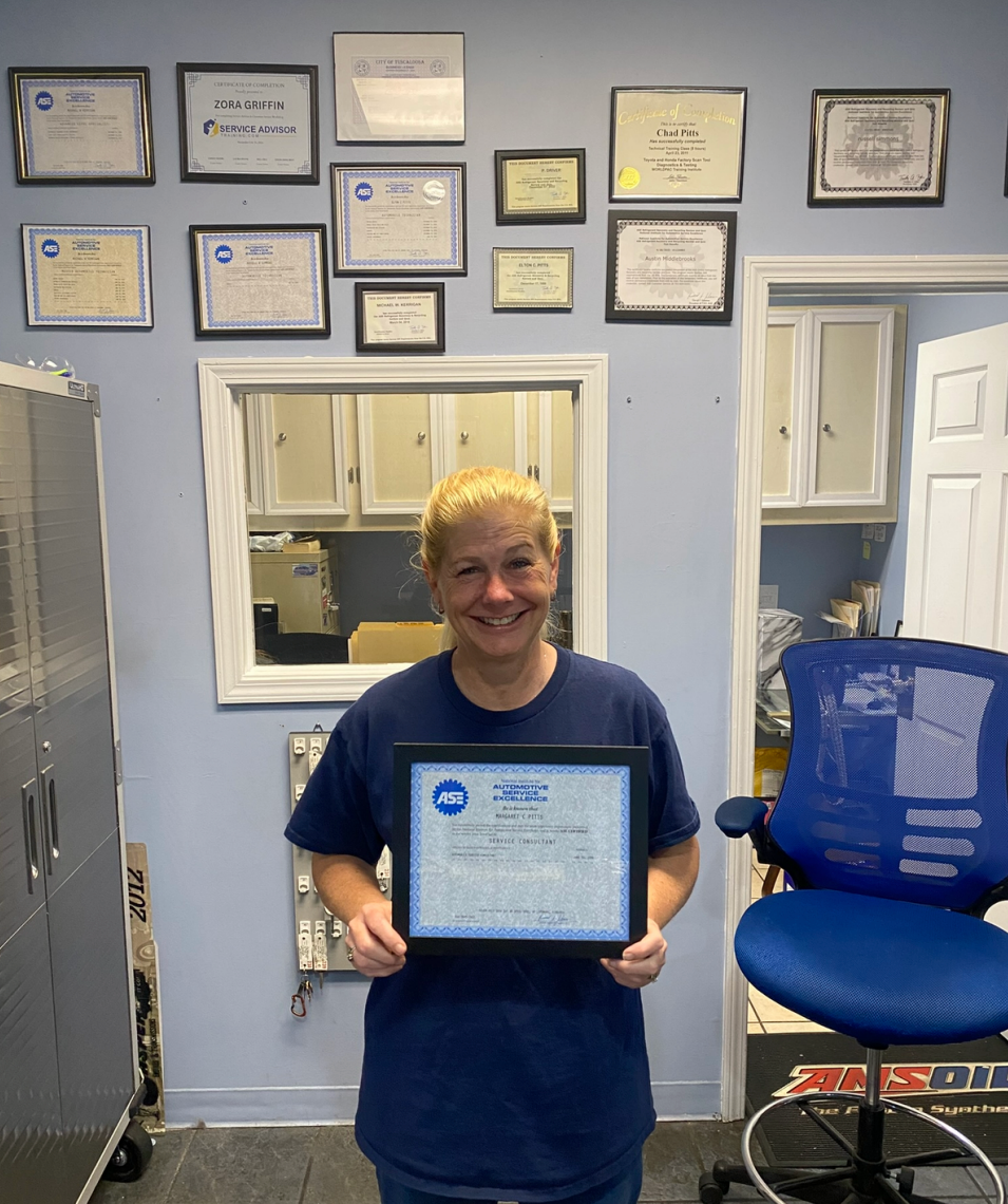 Smiling person holding a framed certificate in an office with awards on the wall. | Pitts Automotive