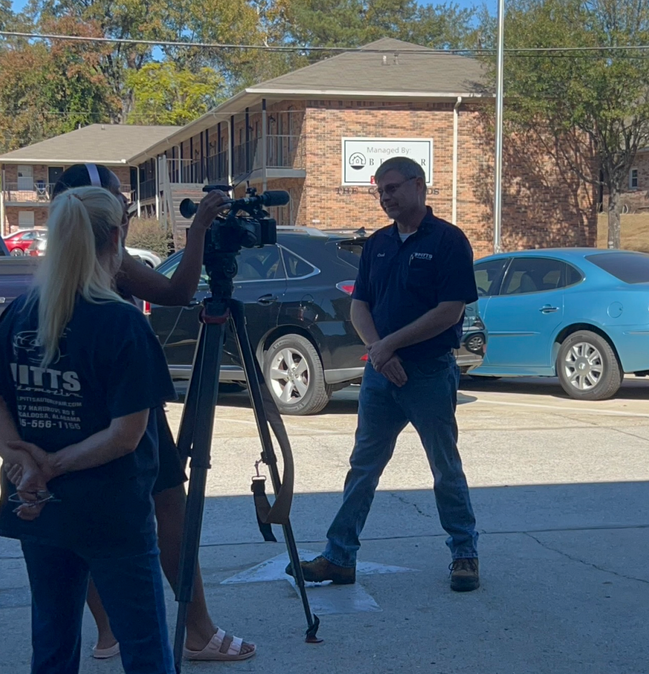 People filming a man with a tripod camera in a parking lot outside a brick building. | Pitts Automotive