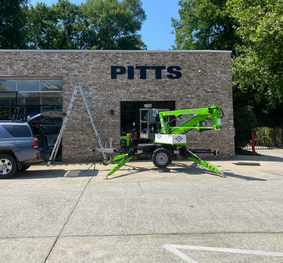 Green cherry picker parked outside a brick building labeled “PITTS” under a sunny sky | Pitts Automotive