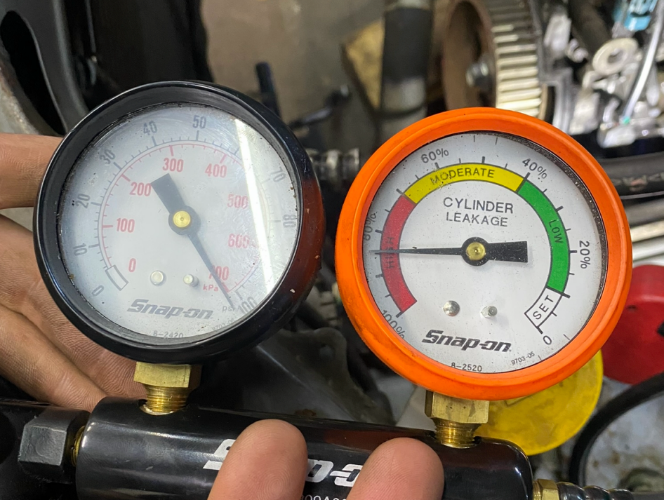 Two Snap-on gauges in a mechanic’s hand, one black and one orange, measuring pressure/ cylinder leakage. | Pitts Automotive