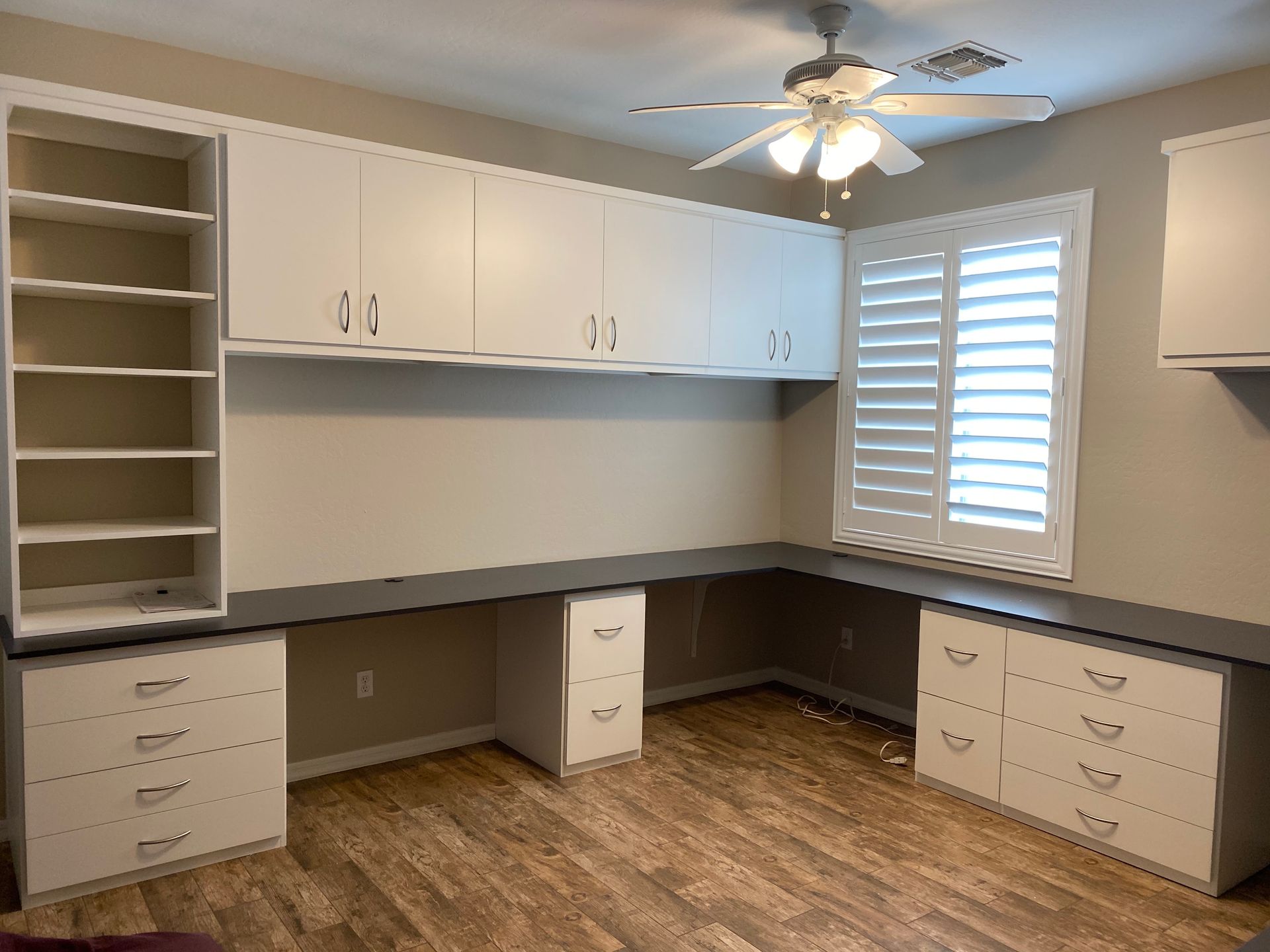 There is a ceiling fan in the home office with a lot of cabinets and drawers.