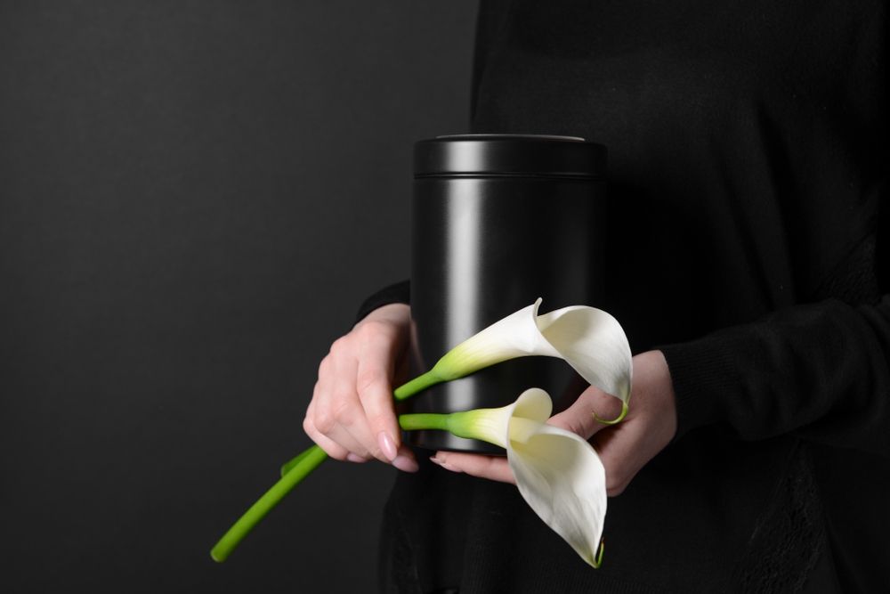 A Woman is Holding a Black Urn With White Flowers in Her Hands — Becker Family Funerals In Forster, NSW
