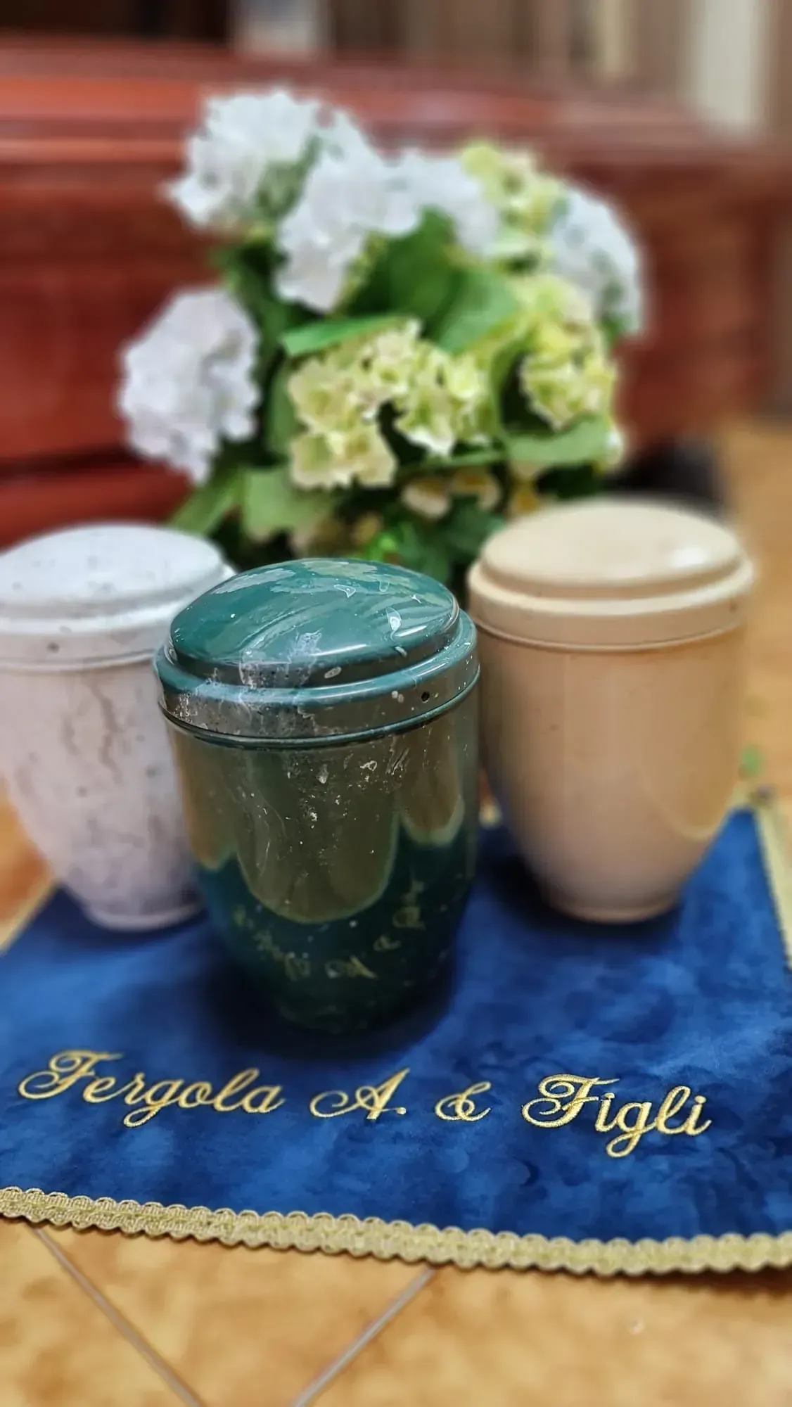 Tre vasi con coperchio su una tovaglia blu, con fiori sullo sfondo e la scritta dorata 