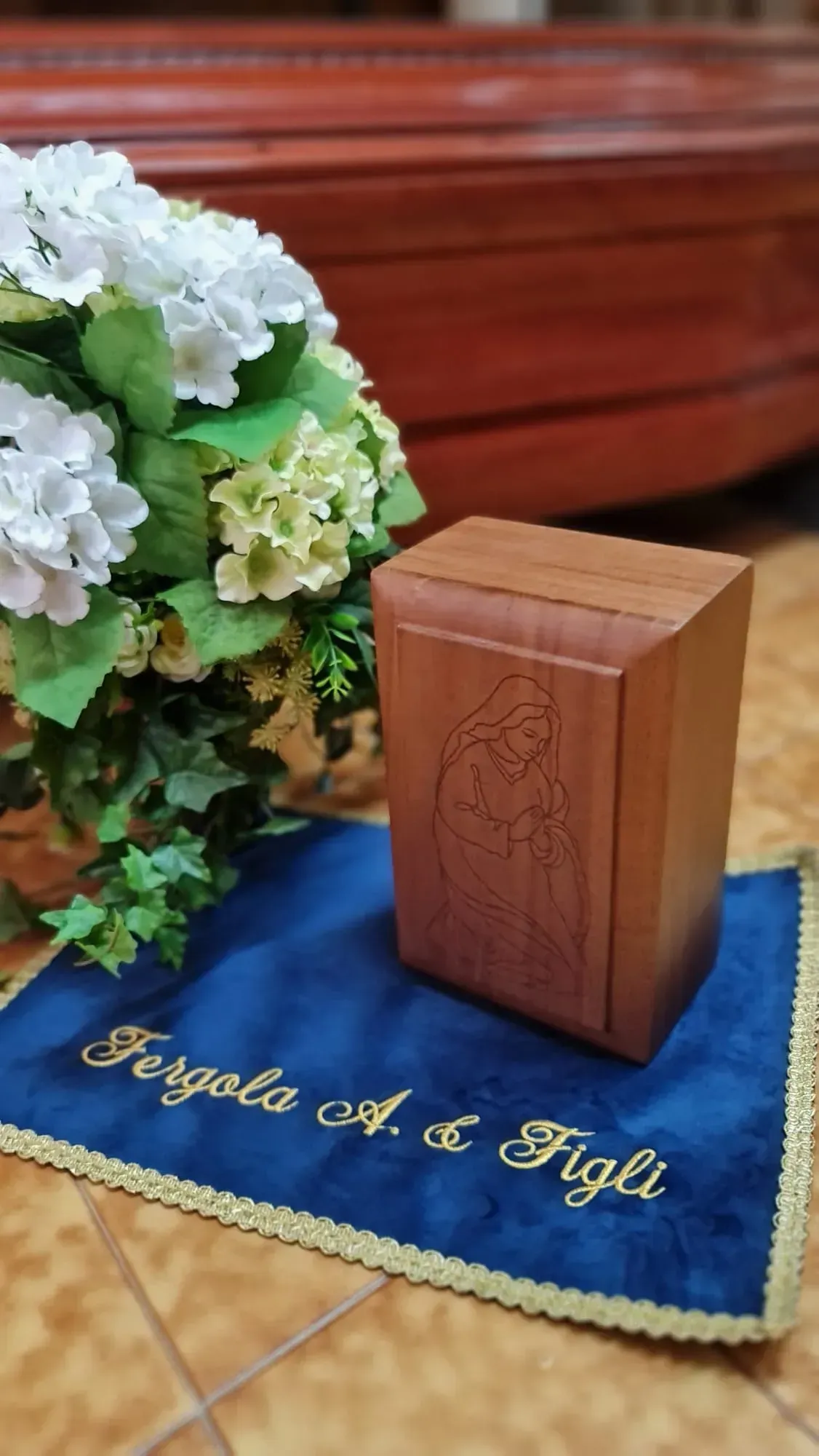 Un'urna di legno su un drappo blu accanto a fiori bianchi e verdi in un ambiente ecclesiastico.