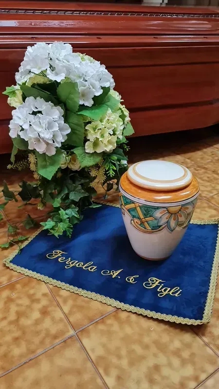 Fiori bianchi e verde accanto a un vaso con coperchio su una tovaglia blu, su un pavimento piastrellato.