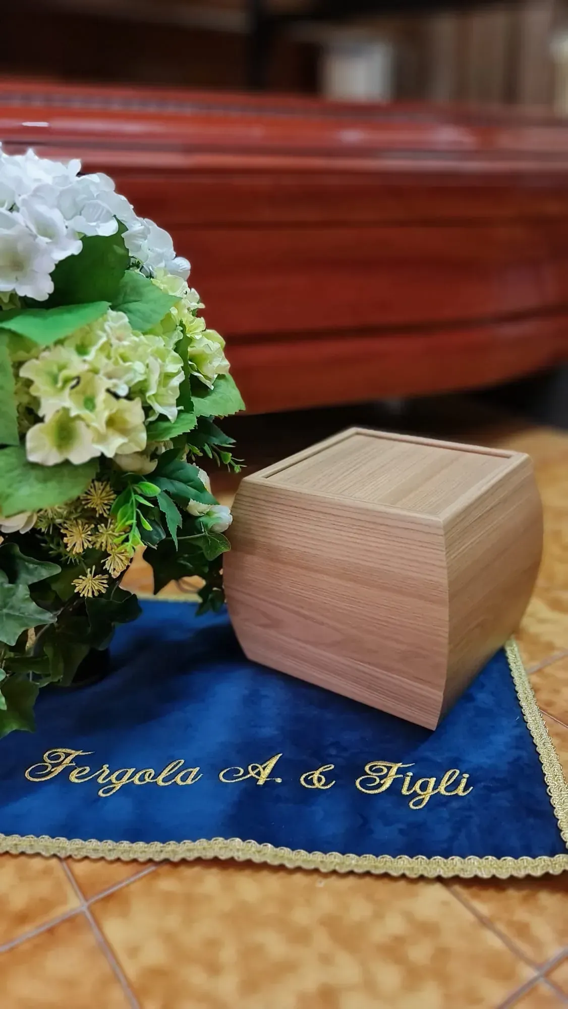 Una piccola scatola di legno accanto a un panno blu e fiori bianco-verdi su un pavimento piastrellato.