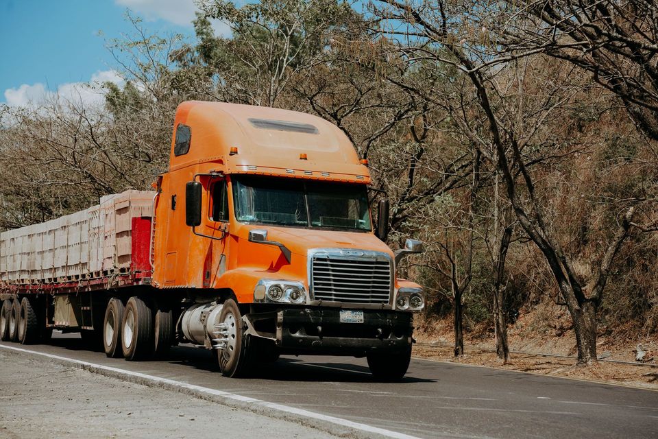 Orange semi-truck driving on a road beside dry trees and brush under a clear sky