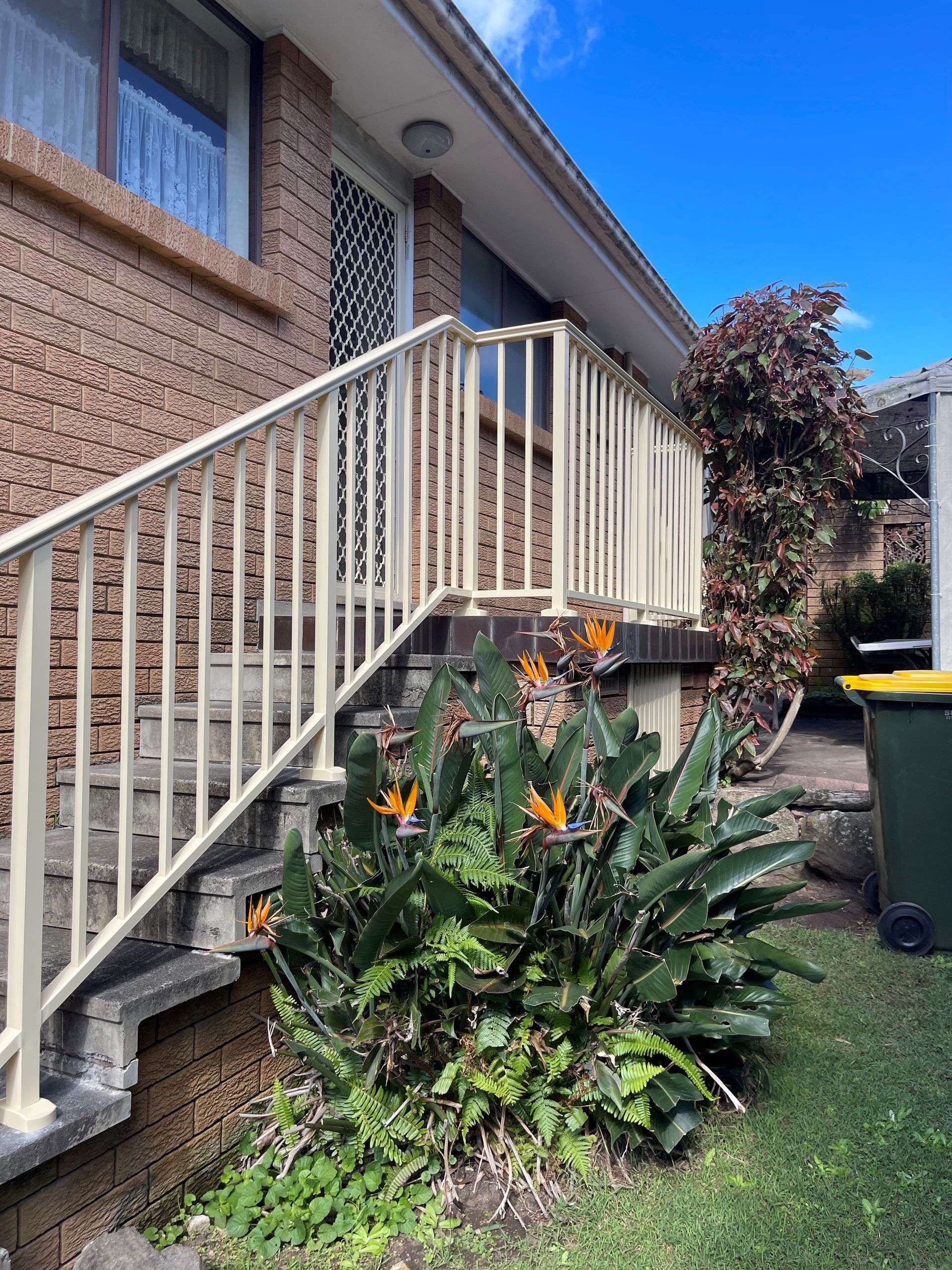 Brick house side entrance with white railing, concrete steps, and orange flowers in front of a green lawn.