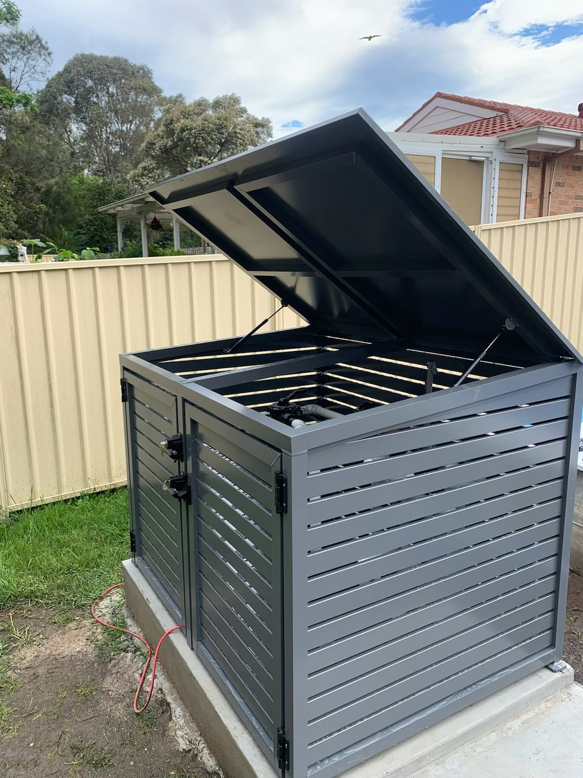 A gray shed with the lid open is sitting next to a fence.