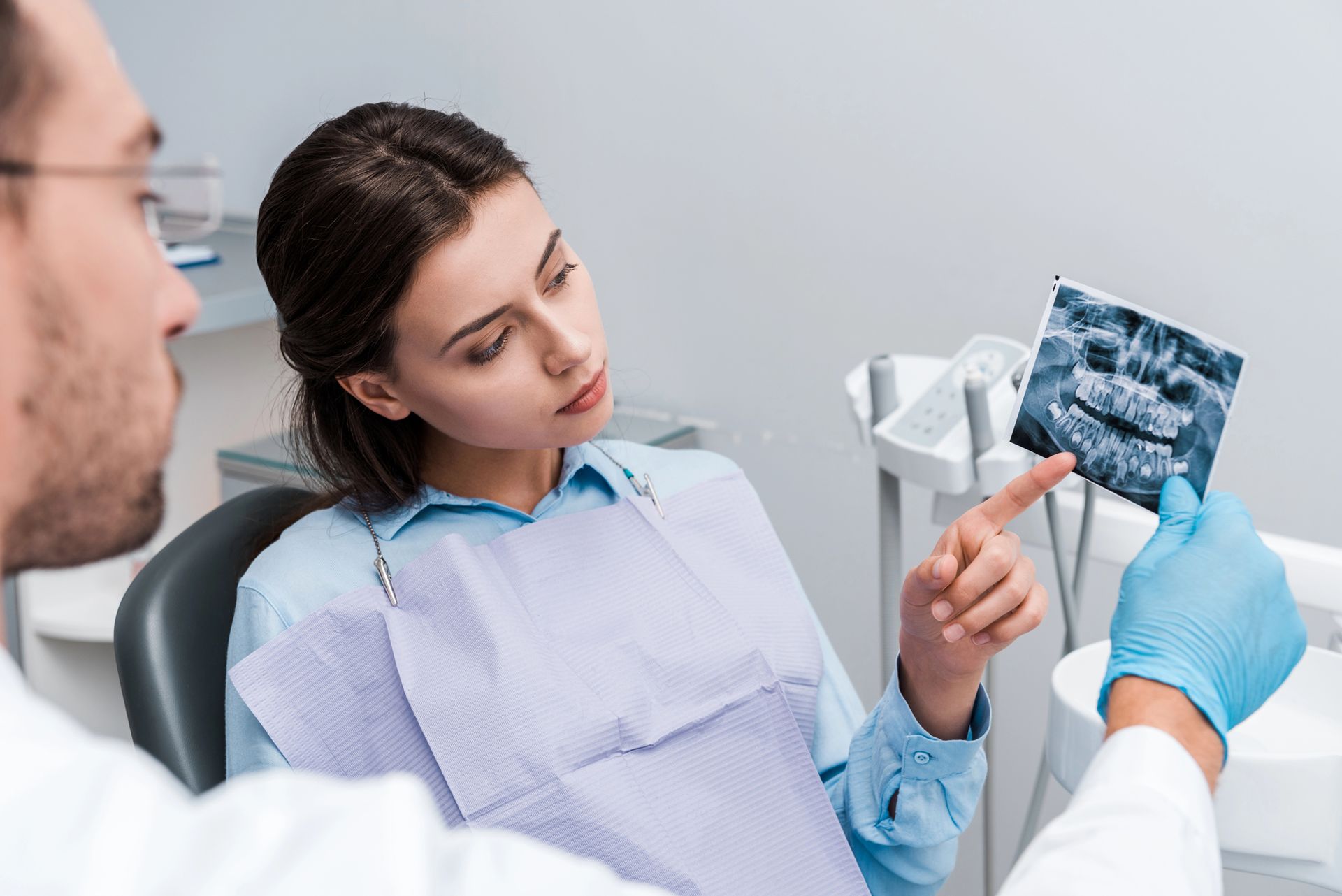 Un dentista con i guanti mostra una radiografia dentale, indicandola mentre ne spiega i risultati a un paziente seduto sulla poltrona odontoiatrica.