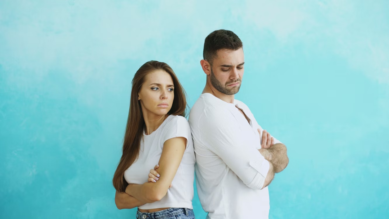 Woman and man standing back-to-back, arms crossed, looking unhappy, against a blue wall.
