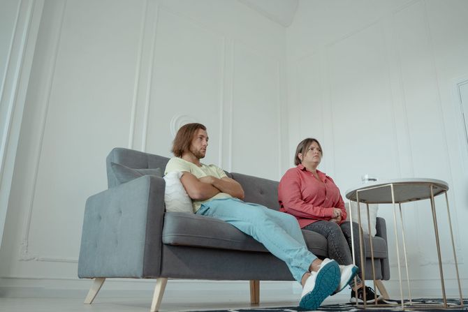 Couple on sofa talking to person holding a clipboard. Man smiles; woman looks on.
