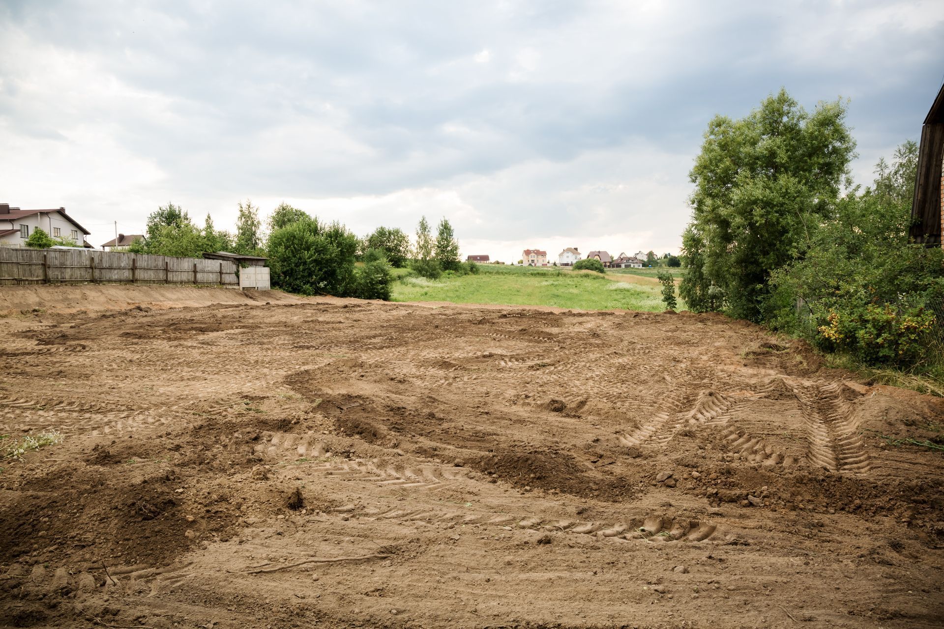 Cleared, leveled dirt lot with tire tracks, under cloudy sky, with houses and trees in the distance.