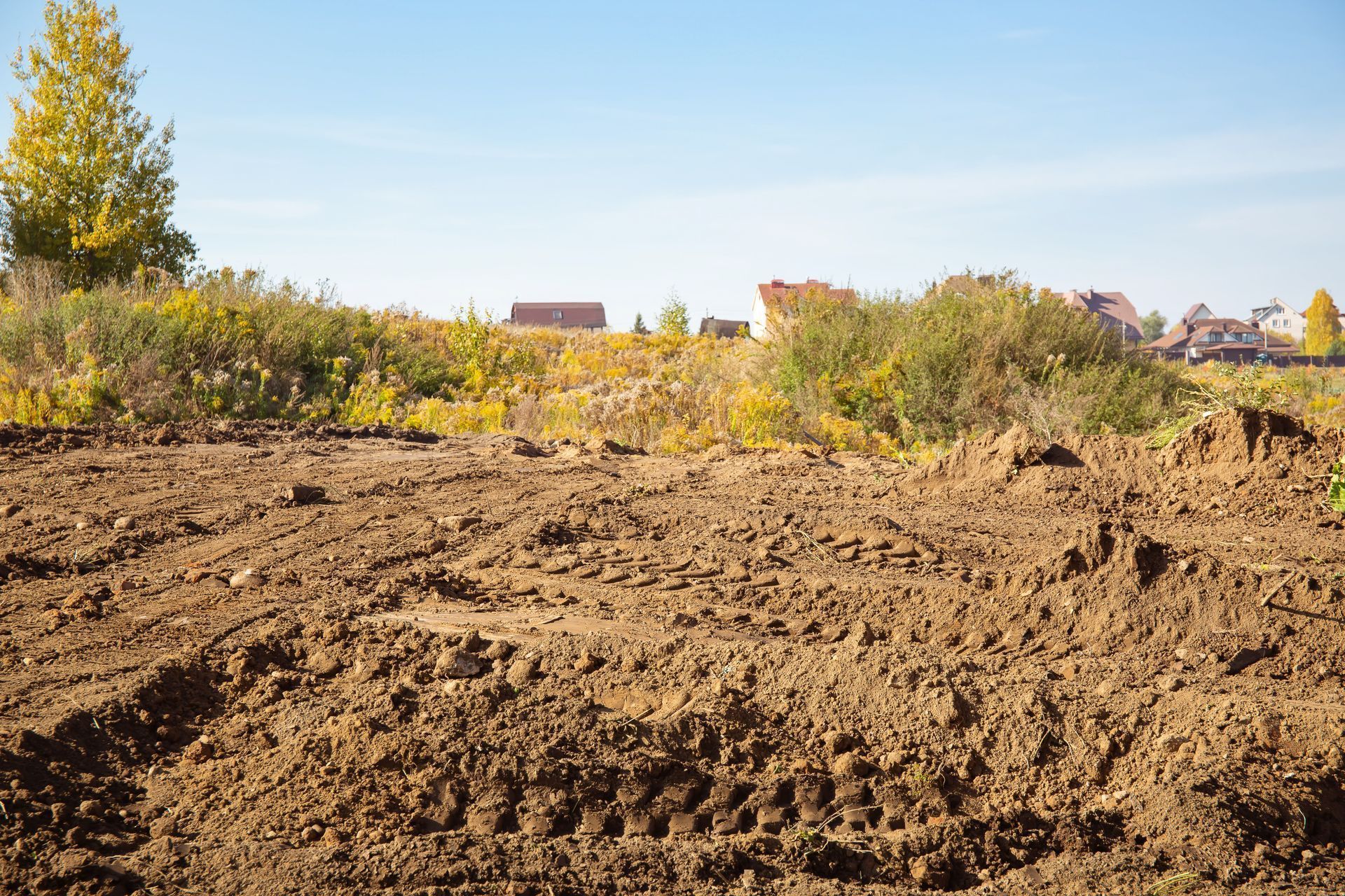 Brown, freshly tilled soil in foreground with vegetation and houses in the distance under a blue sky.