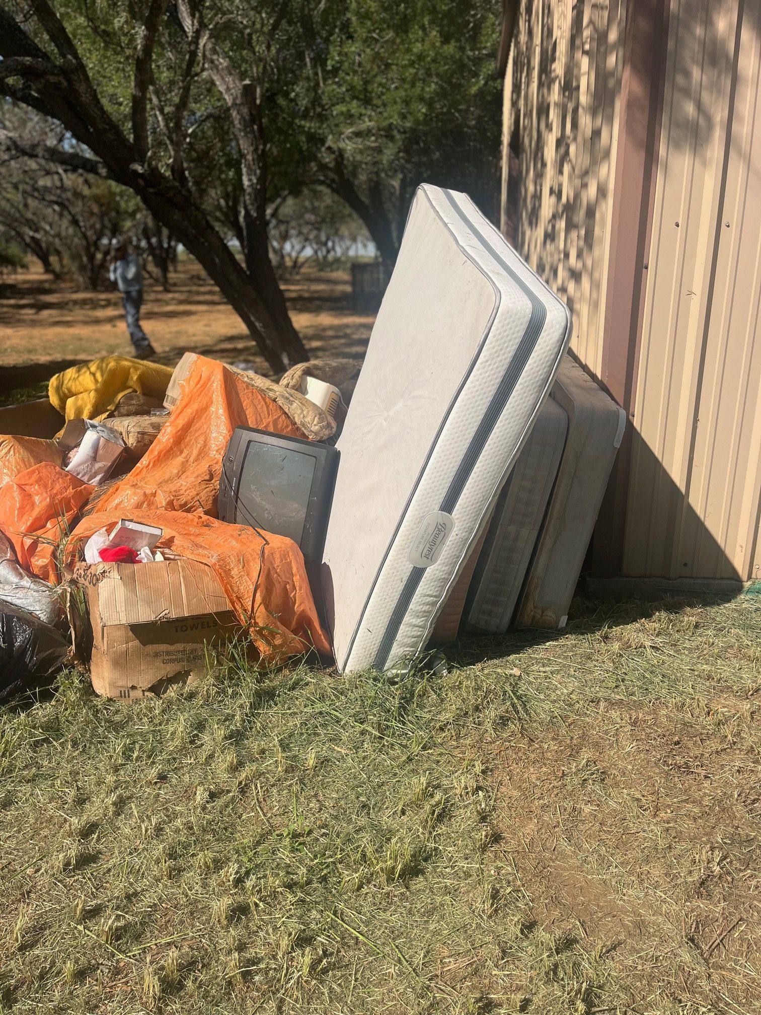 Discarded mattresses and trash piled beside a building under trees, with orange tarps on the ground.