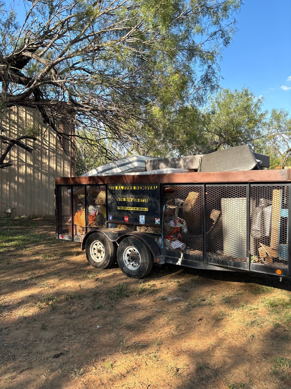Black utility trailer parked under trees beside a house, with equipment inside and a chain-link fence.