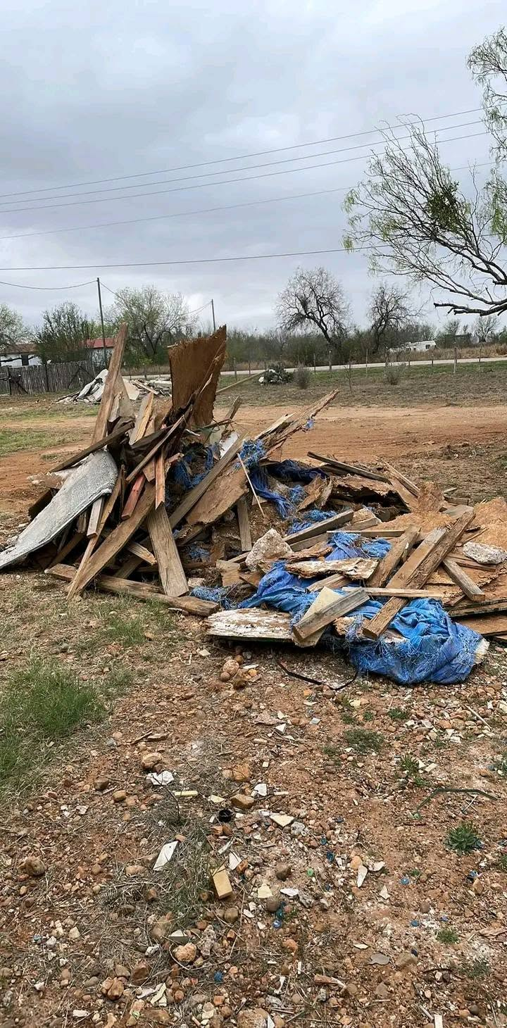 Pile of broken wooden debris and blue tarp on a dirt roadside under a cloudy sky.