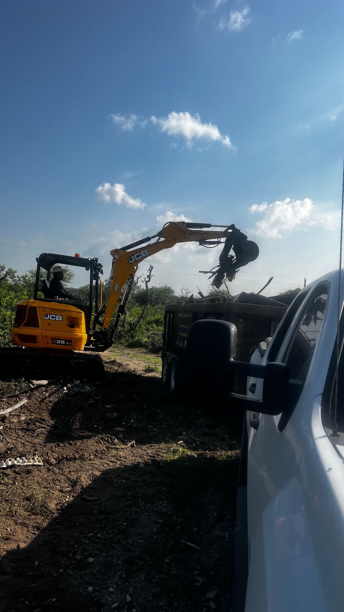 Yellow excavator lifting debris beside a white vehicle under a blue sky.