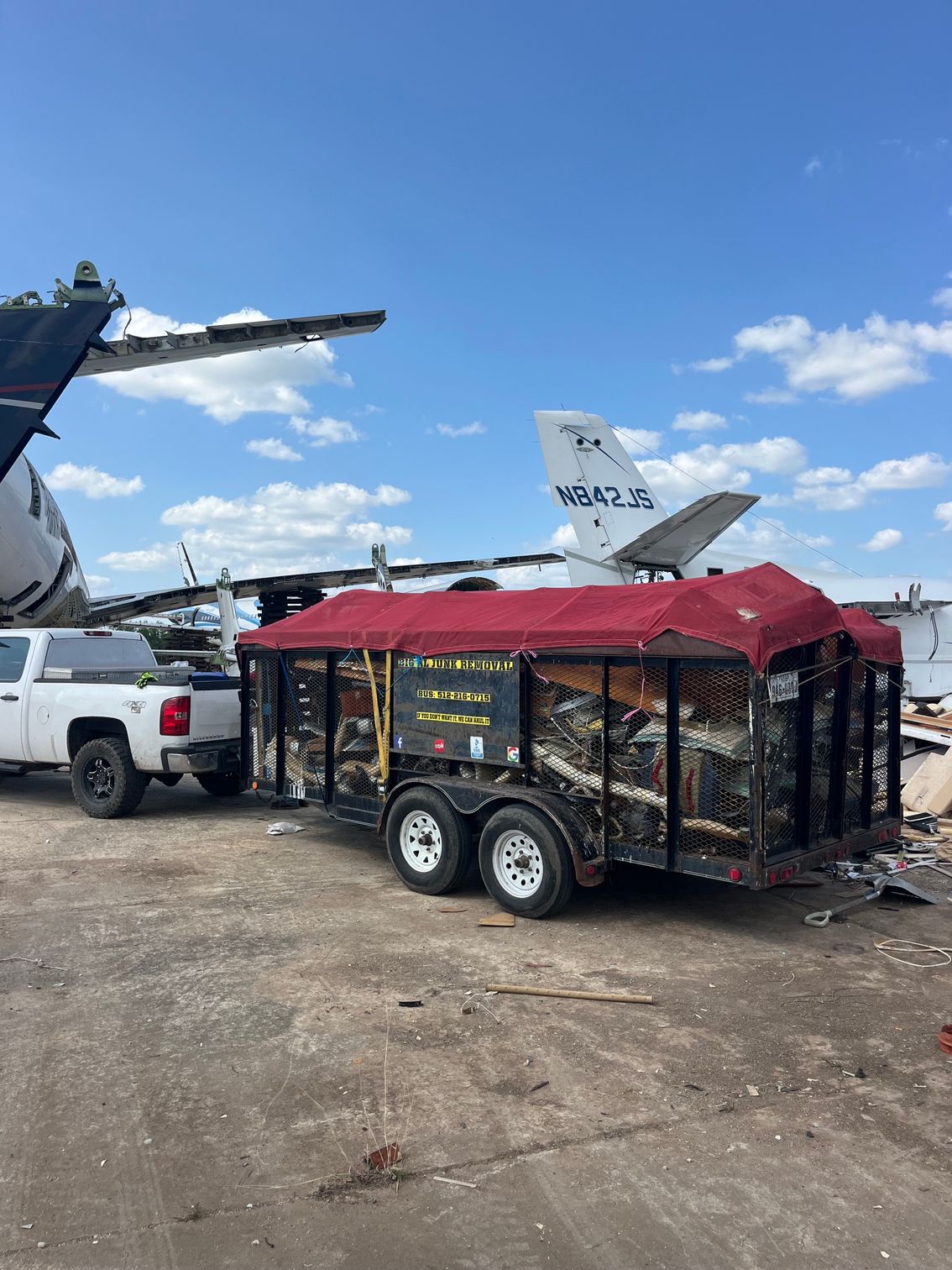 Red utility trailer with black mesh sides parked beside a white pickup and helicopter on a gravel lot under blue sky